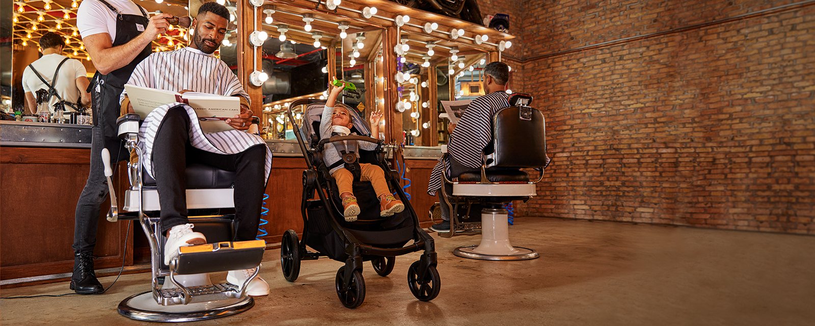 Man in barber chair getting haircut, with child in stroller next to him.