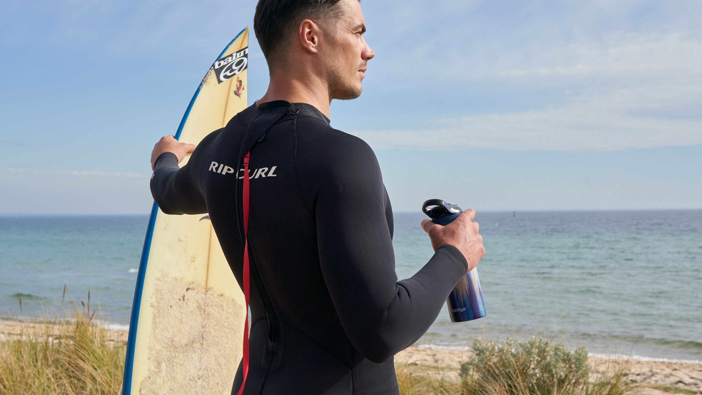 Person holding a surfboard in one hand and a water bottle in the other